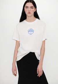 Balts kokvilnas T-krekls ar zilu grafisko apdruku, kurā rakstīts "Moschino Jeans" un "hotels." Īsas piedurknes un brīva piegriezuma, apvienots ar melnu svārku.