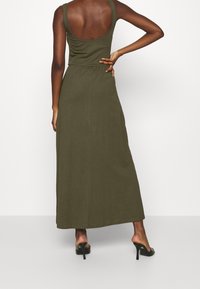 Zielona oliwkowa sukienka maxi z dekoltem z tyłu, dopasowanym gorsetem i elastyczną talią. Gładki materiał, długość do kostek, uzupełniona czarnymi szpilkami.