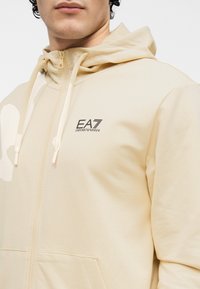 Beige capuchonvest met rits en een gladde textuur, voorzien van een logo op de borst en trekkoorden. Heeft een voorkantzak en een capuchon.