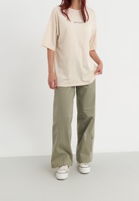 Camiseta de gran tamaño en color beige con texto centrado, combinada con pantalones cargo de pierna ancha en color verde oliva, que cuentan con bolsillos laterales y detalles de costura. Zapatillas deportivas blancas.