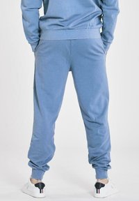 Person wearing blue sweatpants and matching sweatshirt, standing with hands in pockets, white sneakers with black heel detail visible.