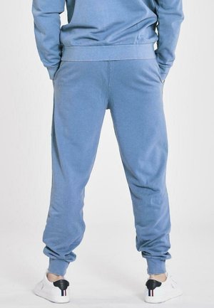 Persona vestida con pantalones de chándal azules y sudadera a juego, de pie con las manos en los bolsillos, zapatillas blancas con detalle negro en el talón visibles.