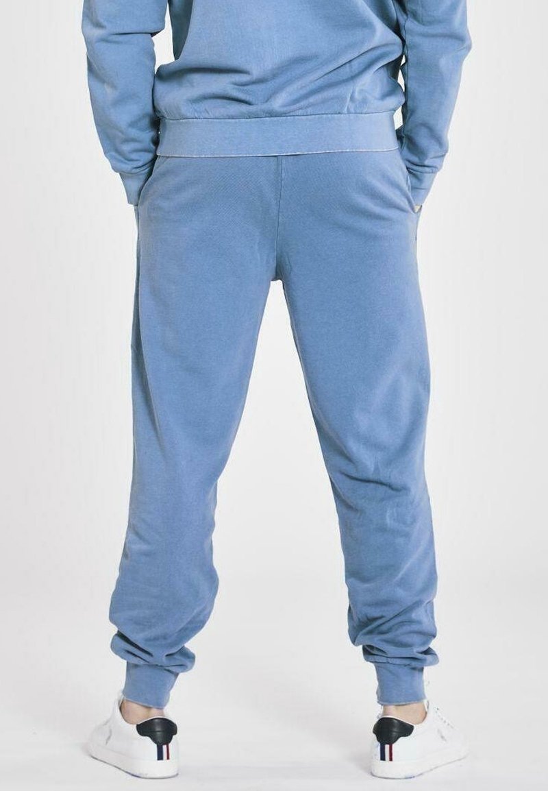 Person wearing blue sweatpants and matching sweatshirt, standing with hands in pockets, white sneakers with black heel detail visible.