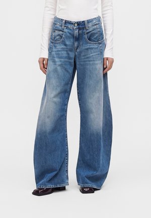 Mannequin portant un jean en denim bleu à jambes larges avec des fentes sur les côtés, un haut blanc à manches longues et des chaussures noires à bout carré.