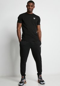 Camiseta negra de manga corta con un pequeño logotipo blanco, combinada con joggers negros que tienen bolsillos laterales. El modelo lleva zapatillas deportivas multicolores.