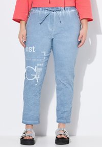 Pantalones de tela arrugada en azul claro con cintura ajustable. Presentan texto impreso y gráficos en la pierna izquierda. Combinados con sandalias plateadas.