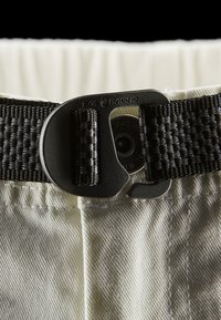 Vit tyg med en texturerad yta pryds av ett svart vävt bälte, säkrat med en metallspänne med en graverad logotyp. Närbild.