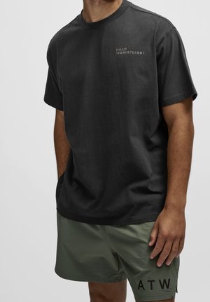 Hombre con camiseta negra de manga corta con el texto "HALO" y números en el pecho, combinada con pantalones cortos verde oliva con la etiqueta "ATW" en la pierna izquierda.