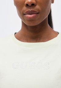 Κοντινό πλάνο ενός ατόμου που φοράει ανοιχτόχρωμο πουκάμισο με τη λέξη «GUESS» σχηματισμένη από μικρές στρας στο στήθος πάνω σε απλό φόντο.