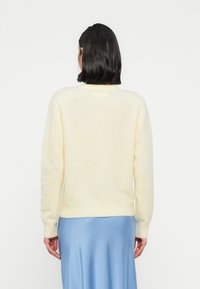 Απαλό κίτρινο πουλόβερ με απαλό ύφασμα και στρογγυλή λαιμόκοψη με ριμπ μανσέτες, εμφανίζεται από πίσω, συνδυασμένο με φούστα σε ανοιχτό μπλε χρώμα.