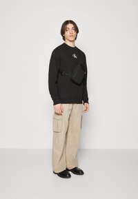 Black sweatshirt with logo, beige cargo pants with large pockets, and black boots. Small black crossbody bag on the right shoulder.