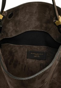 Interieur van een bruine suède handtas met een zwarte voering. Bevat een leren label met de tekst "GIANNI CHIARINI" en gouden hardware accenten.