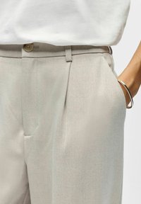 Pantalon beige sur mesure avec une texture lisse, doté d'une fermeture à bouton, de poches latérales et d'un pli unique à l'avant.