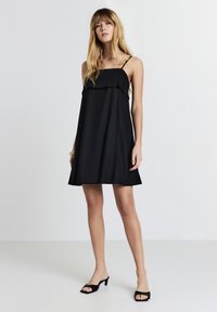 Vestito nero senza maniche con dettaglio a volant stratificato sul corpino, silhouette fluida e spalline regolabili. Indossato con mule con tacco neri.