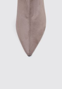 Zapato de punta afilada en suave gamuza color topo. Presenta una costura a lo largo del frente y una textura suave. Las suelas son negras.