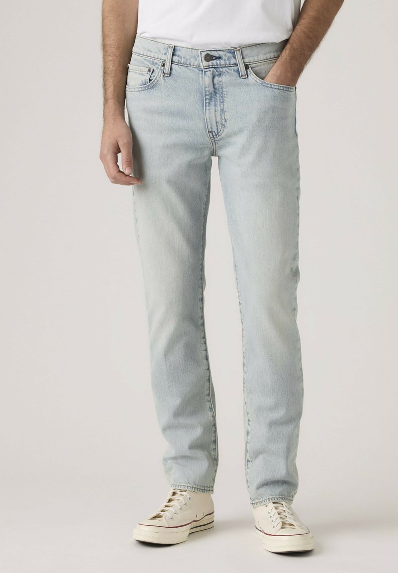 Man wearing light blue straight-leg jeans and white sneakers, with one hand in pocket against plain background.