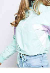 Chaqueta ligera de color verde menta con cremalleras, acentos en púrpura y blanco, puños elásticos y cintura ajustable con cordón. Textura suave.