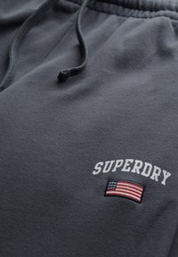 Tissu gris foncé avec une ceinture élastique et un cordon, brodé du texte "SUPERDRY" au-dessus d'un petit écusson drapeau américain.