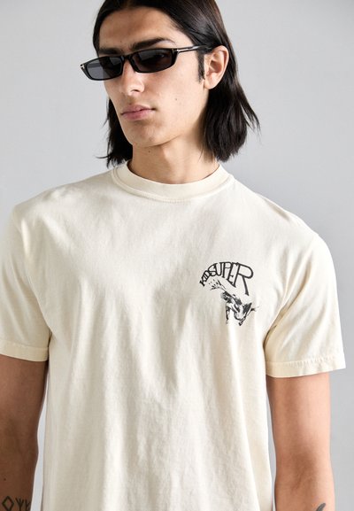 Tricou din bumbac de culoare crem, cu mâneci scurte, decolteu rotund și un imprimeu grafic negru cu cuvântul "KIDSUPER" pe piept.