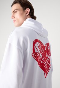 Felpa bianca con cappuccio, caratterizzata da una grande grafica di un cuore rosso sul retro. Tessuto liscio con una vestibilità comoda e cappuccio con coulisse.