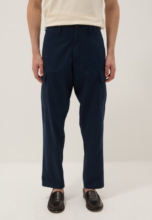 Mann trägt marineblaue Cargohosen und schwarze Loafers, steht vor schlichtem beigem Hintergrund.