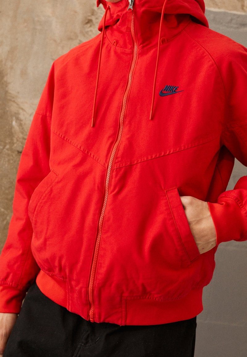 Person som bär en lyseröd Nike zip-up huooded jacka med ena handen i fickan, framför en texturerad beige vägg.