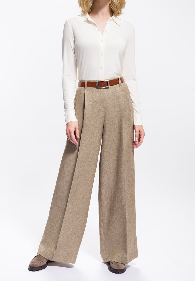 Donna che indossa una camicia bianca a maniche lunghe con bottoni, pantaloni beige a gamba larga, cinta marrone e scarpe taupe, in piedi davanti a uno sfondo bianco.