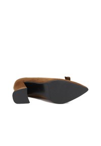 Scarpa con tacco alto in suede marrone, con punta affusolata e tacco basso a blocco. La suola è in gomma nera con una finitura testurizzata. Presenta un fiocco decorativo.