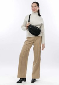 Borsa a tracolla ovale nera con superficie testurizzata e chiusura con zip, indossata sopra un maglione beige a maglia e pantaloni beige a gamba larga. Stivaletti neri con tacco.