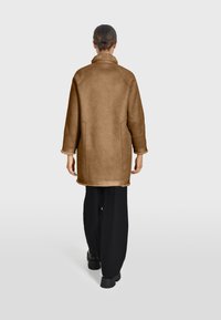 Manteau en shearling marron avec un col montant, des manches longues et des poches latérales, associé à un pantalon large noir et des chaussures noires imposantes.