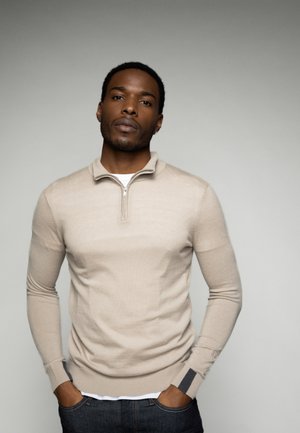 Beige gebreide sweater met een ritskraag, lange mouwen en geribde manchetten met donkere accenten. Draag het over een wit onderhemd en een spijkerbroek.
