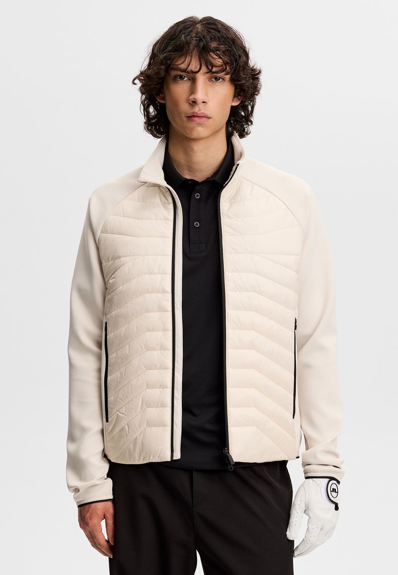 Ung mand med krøllet hår iført en beige quiltet jakke over en sort poloshirt og en hvid golfhandske på højre hånd.