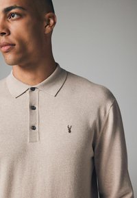 Beige strikket polo med krave, tre knapper og et lille broderet logo på brystet. Glat tekstur og tætsiddende design.