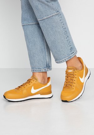 Person wearing mustard yellow Nike sneakers with white swoosh logos and light blue denim jeans standing on a light grey floor.