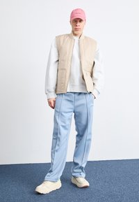 Vesta bej matlasată peste un hanorac gri, asortată cu pantaloni sport albaștri deschiși și o șapcă roz. Adidașii crem completează ținuta.