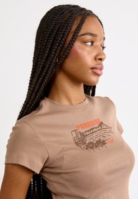 T-shirt en coton marron avec un imprimé graphique d'une scène de plage avec des palmiers et le texte "PARADISE" en orange. Design ajusté à manches courtes.