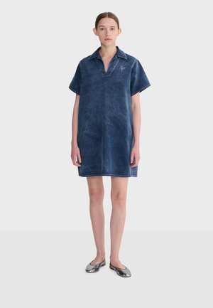 Ung kvinde iført en kortærmet, falmet blå denimkjole og metallisk sølvfarvede flade sko, stående mod en ensartet lys baggrund.