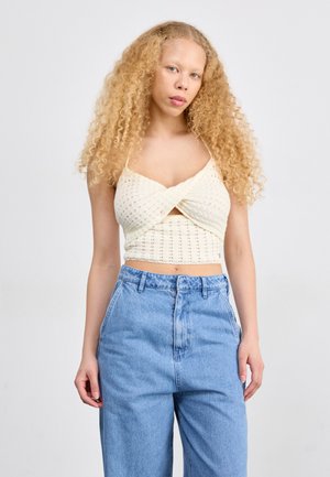 Top corto de punto crema con detalle retorcido en el frente, combinado con jeans de mezclilla de tiro alto de color azul claro. Cabello rizado y fondo neutro.