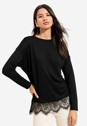 Svart långärmad tröja med rund halsringning och spetskant med blommönster. Mjukt tyg med avslappnad passform, matchat med ljusa byxor.