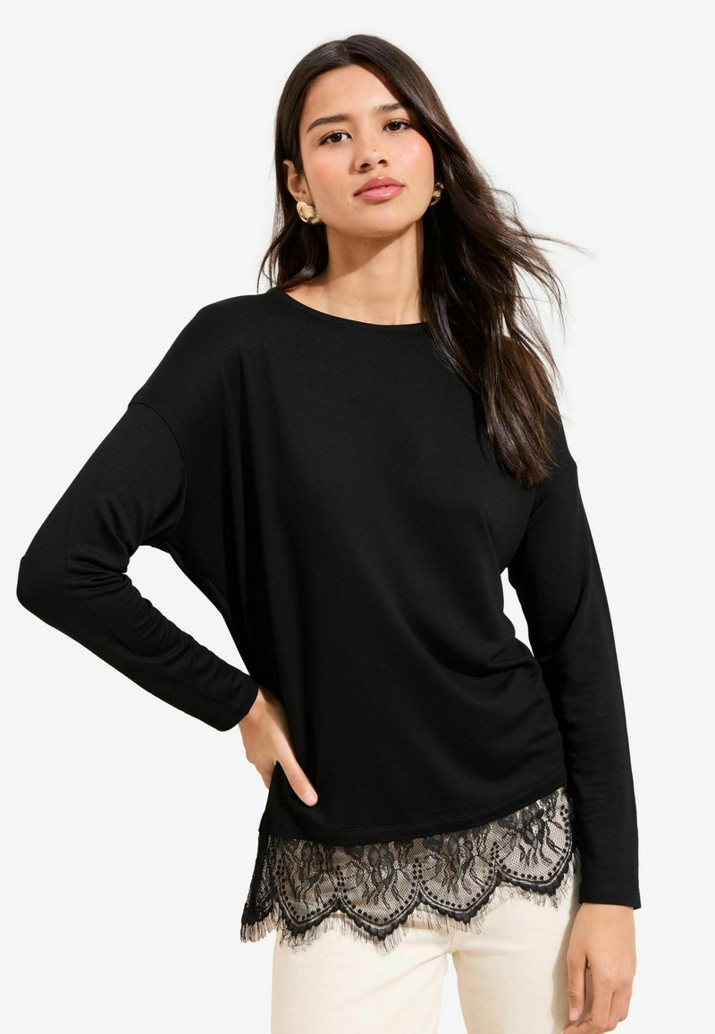 Crna majica s dugim rukavima, okruglim izrezom i čipkanim rubom s cvjetnim uzorkom. Mekani materijal i opušteni kroj, u kombinaciji sa svijetlim hlačama.