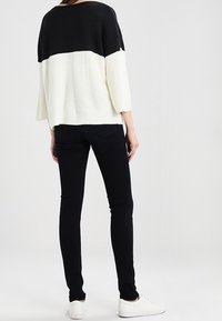 Donna in piedi di spalle che indossa un maglione bianco e nero, jeans skinny neri e scarpe da ginnastica bianche su uno sfondo semplice.
