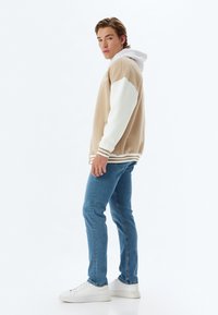 Sudadera con capucha beige y blanca con acentos de rayas, combinada con jeans azules y zapatillas blancas. Ajuste casual con un bolsillo frontal.