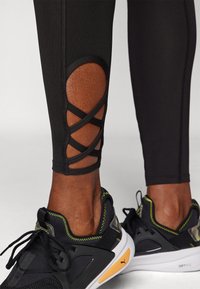 Černé legíny mají výřezový design s křížovými popruhy v oblasti lýtek. Nenosí se s černými sportovními botami se žlutými akcenty a texturovanou látkou.