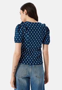 Marineblå top med hvid geometrisk mønster, puffede ærmer, flæser og smocket talje. Sammen med lyseblå jeans.