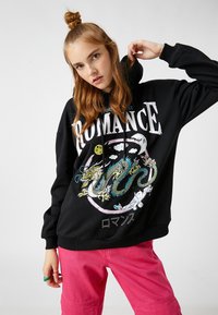 Czarna bluza z kapturem z graficznym wzorem przedstawiającym smoka, chmury oraz słowo "ROMANCE" w białym kolorze. Połączona z różowymi spodniami dla kontrastu.