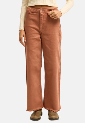 Femme portant un pantalon large couleur rouille avec des ourlets effilochés, des bottes marron clair et un pull court crème, debout devant un fond uni.