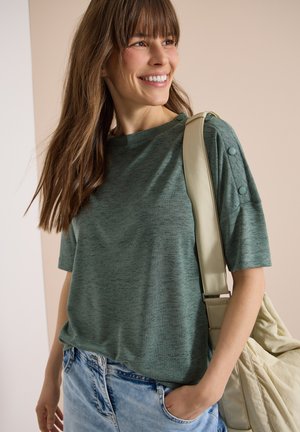Femme souriante aux longs cheveux bruns porte un haut vert à manches courtes avec des détails de boutons et un jean bleu clair, portant un sac beige sur l'épaule.