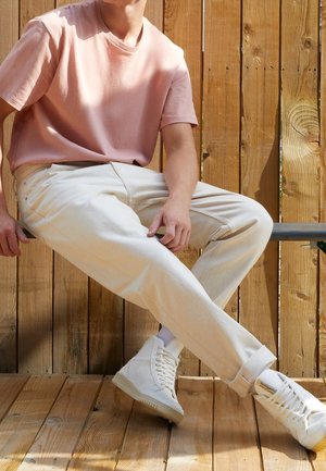 Světle růžová košile s krátkým rukávem, béžové kalhoty a bílé vysoké tenisky. Uvolněný střih s ohrnutými manžetami a dřevěným pozadím.