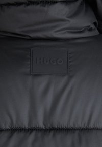 Svart polstret jakke med strukturert stoff, horisontale sømmer og en rektangulær gummilogo med "HUGO" preget.