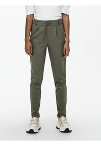 Personne portant un pantalon fuselé vert olive avec taille à cordon et des baskets blanches épaisses, debout devant un fond blanc uni.